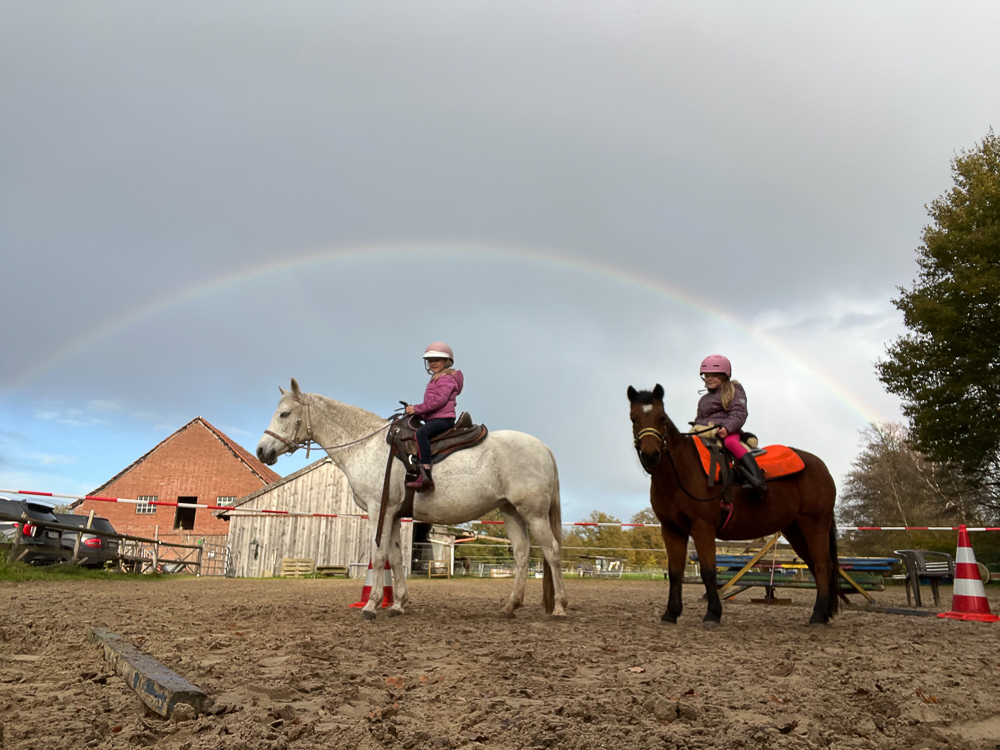 Zwei Kinder sitzen auf Ponys, die auf einem Reitplatz unter einem Regenbogen stehen